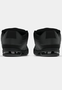 Crne sportske tenisice s podstavljenim ovratnikom, teksturiranim gornjim dijelom i širokom petom s vidljivim jastukom za amortizaciju. Karakteriziraju ih glatke površine i minimalni ukrasi.