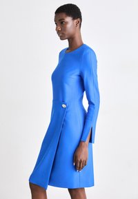 Μπλε φόρεμα με μακριά μανίκια, εφαρμοστό κορσέ και φουσκωτό φουστάνι. Διαθέτει λεπτομέρεια με κουμπί στο πλάι και κοπή στο μανίκι. Λείου υφάσματος.