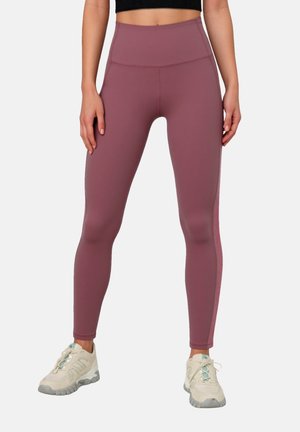 Mujer con leggings malva de cintura alta y zapatos deportivos blancos, de pie con las manos relajadas a los lados contra un fondo liso.