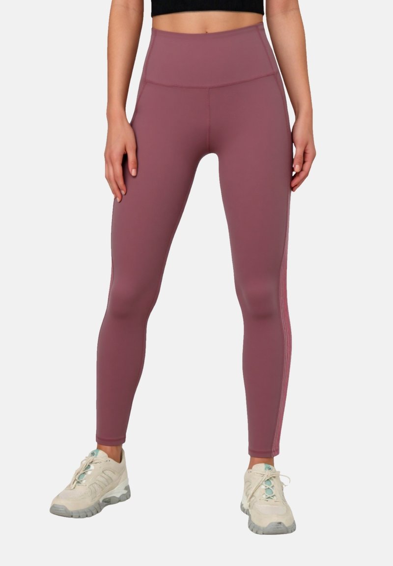 Mujer con leggings malva de cintura alta y zapatos deportivos blancos, de pie con las manos relajadas a los lados contra un fondo liso.