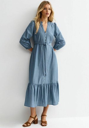 Mujer de pie contra una pared, vistiendo un vestido midi azul de manga larga, abotonado, con cinturón en la cintura y sandalias marrones de tacón ancho.