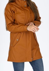 Femme portant une veste à capuche marron avec des poches à boutons-pression et des poignets, associée à un jean bleu, ajustant sa manche sur fond blanc.