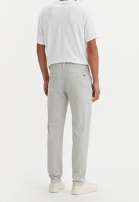 Man wearing light gray pants, white short-sleeve t-shirt, and off-white sneakers, standing against a plain white background, back view.