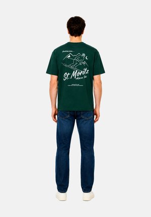 Mann trägt grünes St. Moritz Ski Grafik-T-Shirt, blaue Jeans und weiße Schuhe, steht mit dem Rücken zum Betrachter vor einfachem hellem Hintergrund.
