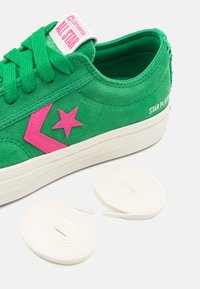Baskets Converse Star Player en daim vert avec logo rose, semelle blanche, étiquette "All Star" et deux lacets blancs enroulés sur fond blanc.