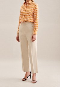 Orange mönstrad skjorta med knappning och krage samt långa ärmar, kombinerad med beigea byxor med hög midja och svarta sandaler med remmar.