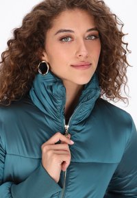 Miętowa kurtka puchowa z wysokim kołnierzem, z zamkiem błyskawicznym i ściągaczami przy rękawach. Modelka ma kręcone włosy i nosi kolczyki z złotymi akcentami.