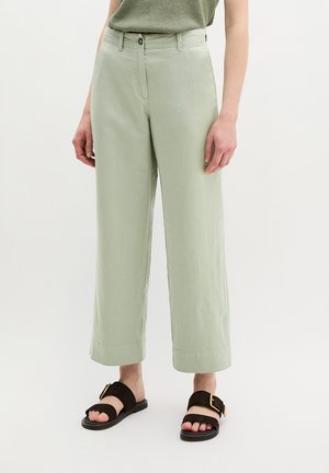 Personne portant un pantalon large court de couleur sauge claire associé à des sandales plates noires avec de grosses boucles dorées.