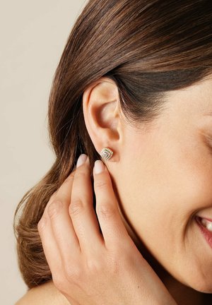 Frau mit braunen Haaren, die einen goldenen, mit Diamanten besetzten quadratischen Ohrstecker berührt, lächelt mit sichtbaren Zähnen.