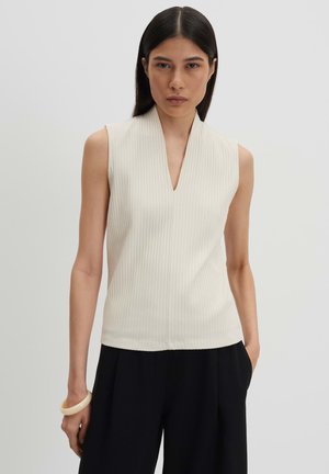 Femme aux cheveux lisses et foncés portant un haut blanc sans manches en côte avec un col en V et un pantalon noir, debout devant un fond clair uni.