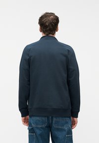 Άνδρας που στέκεται με την πλάτη, φορώντας μπλε ναυτικό φούτερ και μπλε τζιν με πολλές πίσω τσέπες και θηλιές.