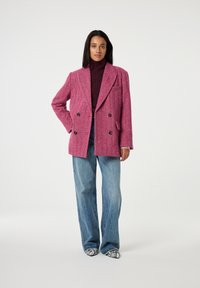 Dubbel-breasted, magenta gestructureerde jas met brede revers, zwarte knopen en twee voordetjes, gecombineerd met lichtblauwe wijde jeans en gemusterde schoenen.