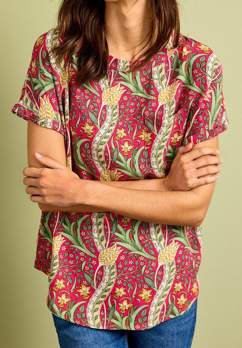 Femme portant un chemisier rouge à manches courtes à motif floral avec des touches vertes et jaunes, bras croisés, sur un fond vert clair.
