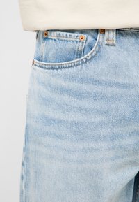 Valge topiga sisse torgatud helesiniste teksade lähedalt vaadatav tasku, mida riivivad kuparist neetide detailid.