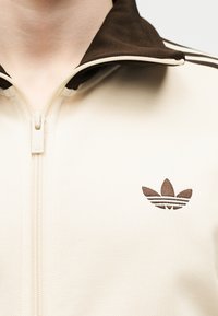Detailný záber na béžovú bundu na zips s tmavohnedým golierom a vyšitým logom Adidas trefoil na hrudi.