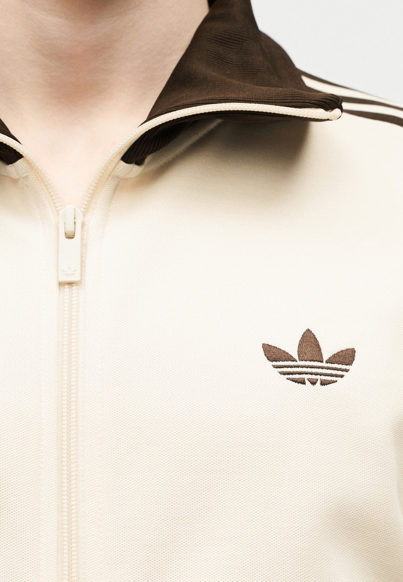 Primer plano de chaqueta beige con cremallera, cuello marrón oscuro y logo trefoil de Adidas bordado en el pecho.