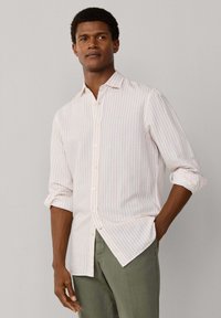 Mies, joka pukeutunut vaaleanpunaiseen ja valkoiseen raitapaita, hihat käännettynä ylös ja yllä oli oliivinvihreät housut, seisoo yksinkertaisen taustan edessä.