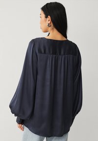 Marinblå satinbluse med lange puffærmer, rund halsudskæring og samlet rygdetalje. Blød tekstur og svævende design.