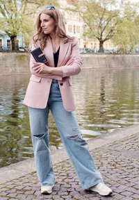 Světle růžový blazér přes černý top, doplněný světle modrými roztrhanými džíny. Bílé tenisky, držící černou tečkovanou peněženku, stojící u vody.