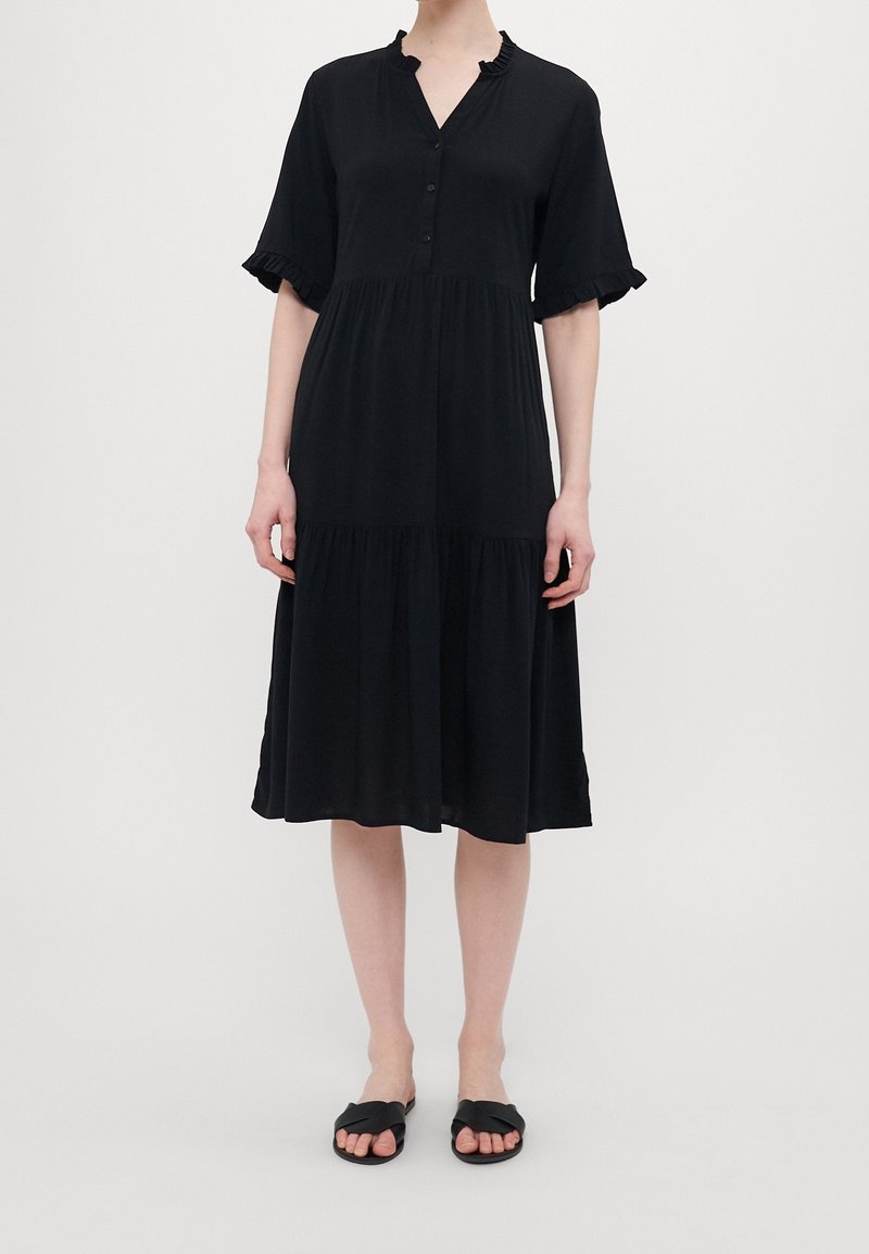Femme portant une robe noire mi-genoux avec des manches courtes à volants, boutonnée à l'avant, et des sandales noires slip-on sur un fond blanc.