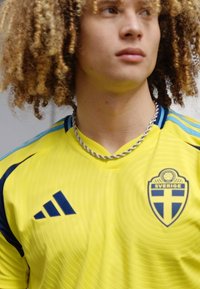 Żółta koszulka Adidas z niebieskimi akcentami i herbem Szwecji, z teksturowanym wzorem i okrągłym dekoltem. Widoczny srebrny naszyjnik.