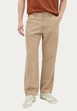 Person iført beige bukser med lige ben og hvide sneakers, med en hånd i lommen og en brun skjorte delvist synlig.