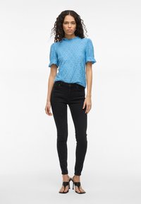 Lyseblå bluse med hullmønster, rufflekrage og -ermer, kombinert med svarte smale jeans og svarte hælsandaler.