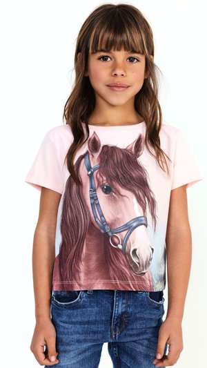 Ragazza giovane con capelli castani di media lunghezza, che indossa una maglietta rosa con un'illustrazione dettagliata del volto di un cavallo e jeans blu.