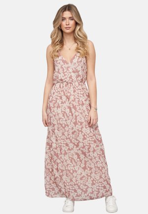 Kvinde med langt bølget hår iført en ærmeløs pink maxikjole med hvidt blomsterprint og hvide sneakers, stående mod en ensfarvet baggrund.