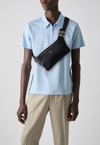 Bandolera de nylon negra llevada sobre una camiseta polo azul claro. La bolsa presenta un logo triangular, un bolsillo con cremallera y una correa ajustable.