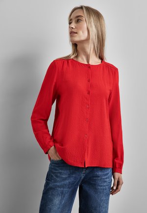 Blusa rossa a maniche lunghe in tessuto strutturato, con scollatura rotonda e chiusura con bottoni. Abbinata a jeans in denim blu, dal fit casual.
