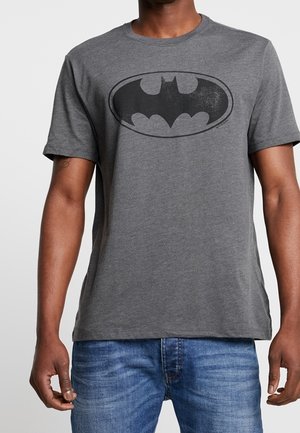 Osoba ubrana w szarą koszulkę z czarnym logo Batmana oraz niebieskie dżinsy, stojąca na jednolitym jasnym tle.