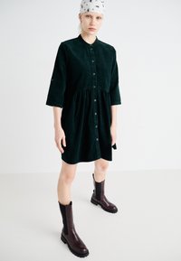 Robe en velours côtelé vert foncé avec un devant à boutons, une taille élastique et des manches trois-quarts, assortie à des bottes marron jusqu'aux genoux.