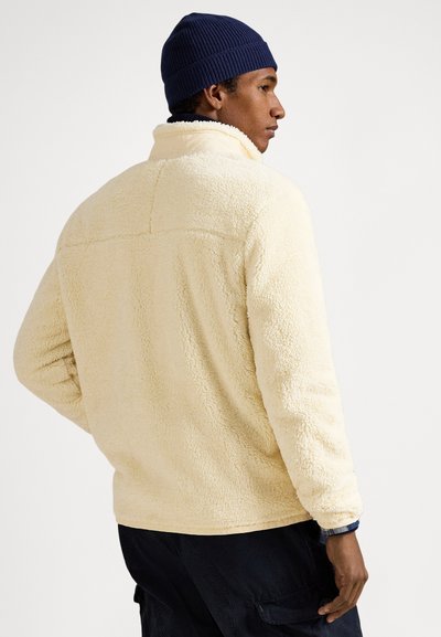 Άνδρας που φοράει κρέμ φλις μπουφάν, σκούφο ναυτικού μπλε και σκούρο παντελόνι, στέκεται με πλάτη και προφίλ πλευράς ορατά σε λευκό φόντο.