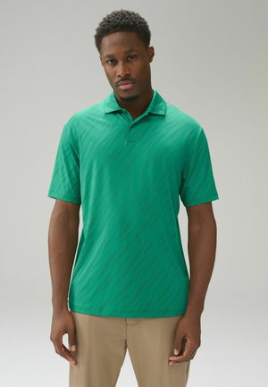 Mann in grünem Kurzarm-Poloshirt und beigen Hosen steht vor einem schlichten, hellen Hintergrund, schaut geradeaus mit neutralem Gesichtsausdruck.