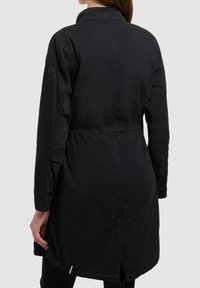 Nő hosszú fekete kabátban, amely deréknál meg van húzva, háttal áll egy egyszerű világos háttér előtt.