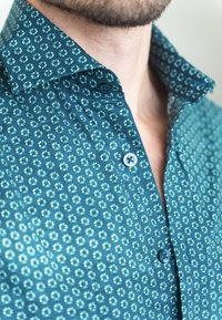 Mann trägt ein türkisfarbenes Hemd mit kleinem weißem Blumenmuster und drei sichtbaren Knöpfen, Kragen offen, zeigt Hals und Kinnbart.