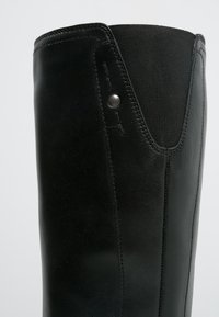 Botte en cuir noir avec une finition lisse, un panneau extensible et un détail à pression métallique près de la cheville. Le design est haut et ajusté.