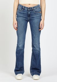 Frau in blauen Bootcut-Jeans und weißen Turnschuhen, die vor einem schlichten hellen Hintergrund steht.