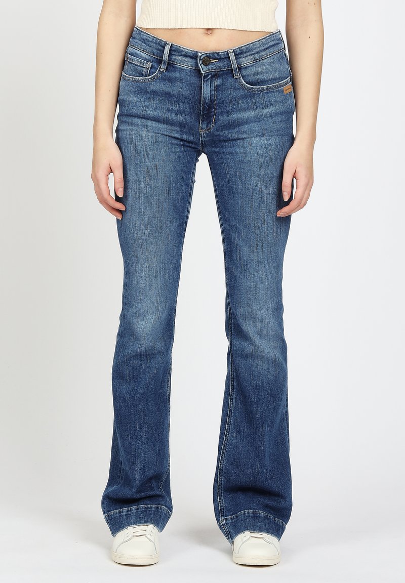 Frau in blauen Bootcut-Jeans und weißen Turnschuhen, die vor einem schlichten hellen Hintergrund steht.