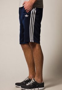 Navy blue athletic shorts with white side stripes and an Adidas logo. worn with black sneakers featuring white laces and textured soles.