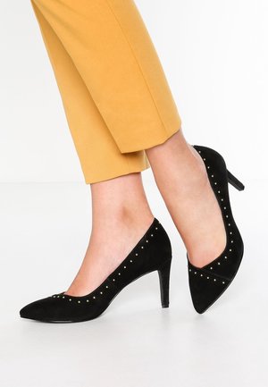 Pieds portant un pantalon jaune moutarde et des escarpins en daim noir à bout pointu, ornés de petits clous dorés et talons aiguille de hauteur moyenne.