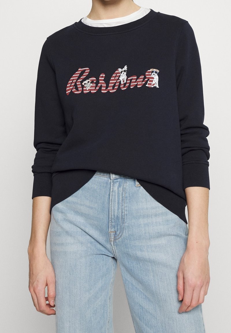 Personne portant un sweat-shirt noir avec le texte rayé « Barbour » et des illustrations de chiens, associé à un jean bleu clair.