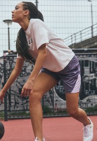 Sportkleding met een wit t-shirt met logo en paarse Nike-shorts. Scène gevestigd op een roze baan te midden van een met graffiti bedekte achtergrond.