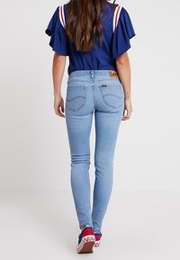 Jeans ajustados de color azul con cintura alta, que cuentan con una etiqueta de la marca Lee, diseños sutiles en los bolsillos traseros y combinados con zapatillas rojas y azules.