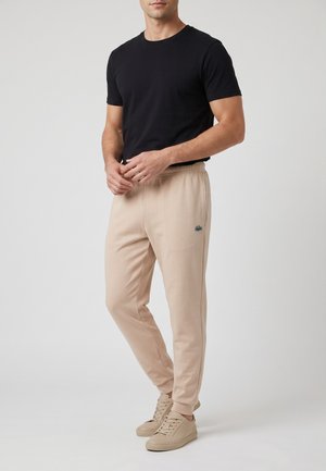 Uomo con t-shirt nera a maniche corte, pantaloni da jogging beige con logo e scarpe da ginnastica beige, in piedi davanti a uno sfondo semplice.