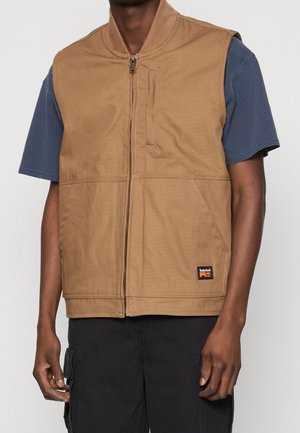 Homme portant un gilet de travail beige zippé sur une chemise bleu marine à manches courtes et un pantalon cargo noir, debout sur un fond uni clair.