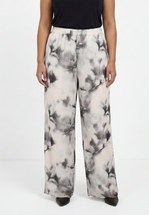 Femme portant un pantalon à jambes larges avec un motif floral abstrait noir et blanc et des chaussures noires pointues sur un fond uni.