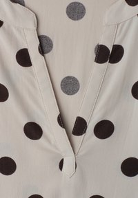 Tissu crème avec de gros pois noirs et gris et un détail d'encolure cousu en forme de V.