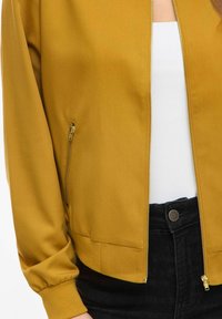 Veste bomber jaune moutarde à texture lisse, fermeture éclair à l'avant, poignets et ceinture côtelés. Comprend des poches latérales avec fermetures éclair dorées.
